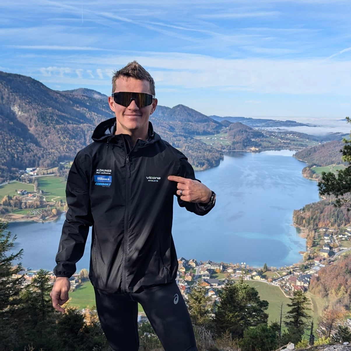 Bergläufer in Sportbekleidung steht auf einem Berg und ein See ist im Hintergrund zu sehen.